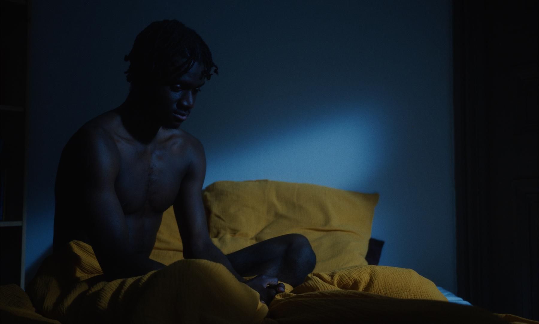 Photogramme : un jeune homme assis sur le lit jaune, dans une lumière bleue de nuit.