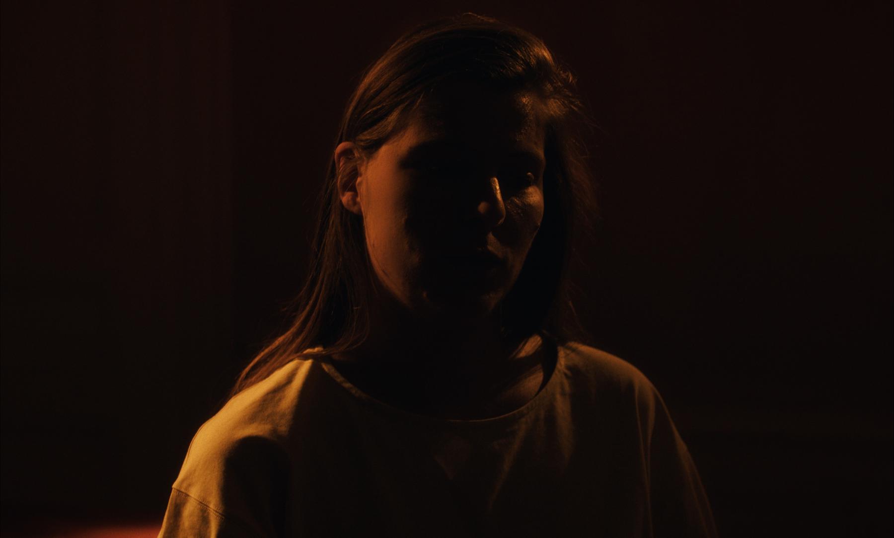 Photogramme : une femme dans la pénombre orangée, le visage à demi éclairé.