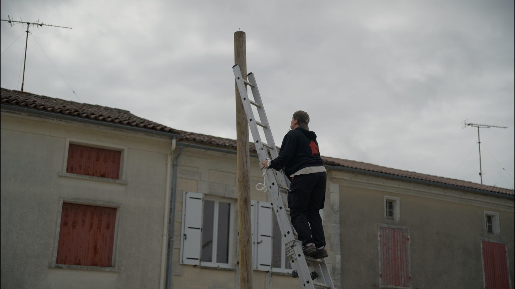 Lieu Dire : un bénévole en haut d'une échelle installe un mât devant les maisons du village.