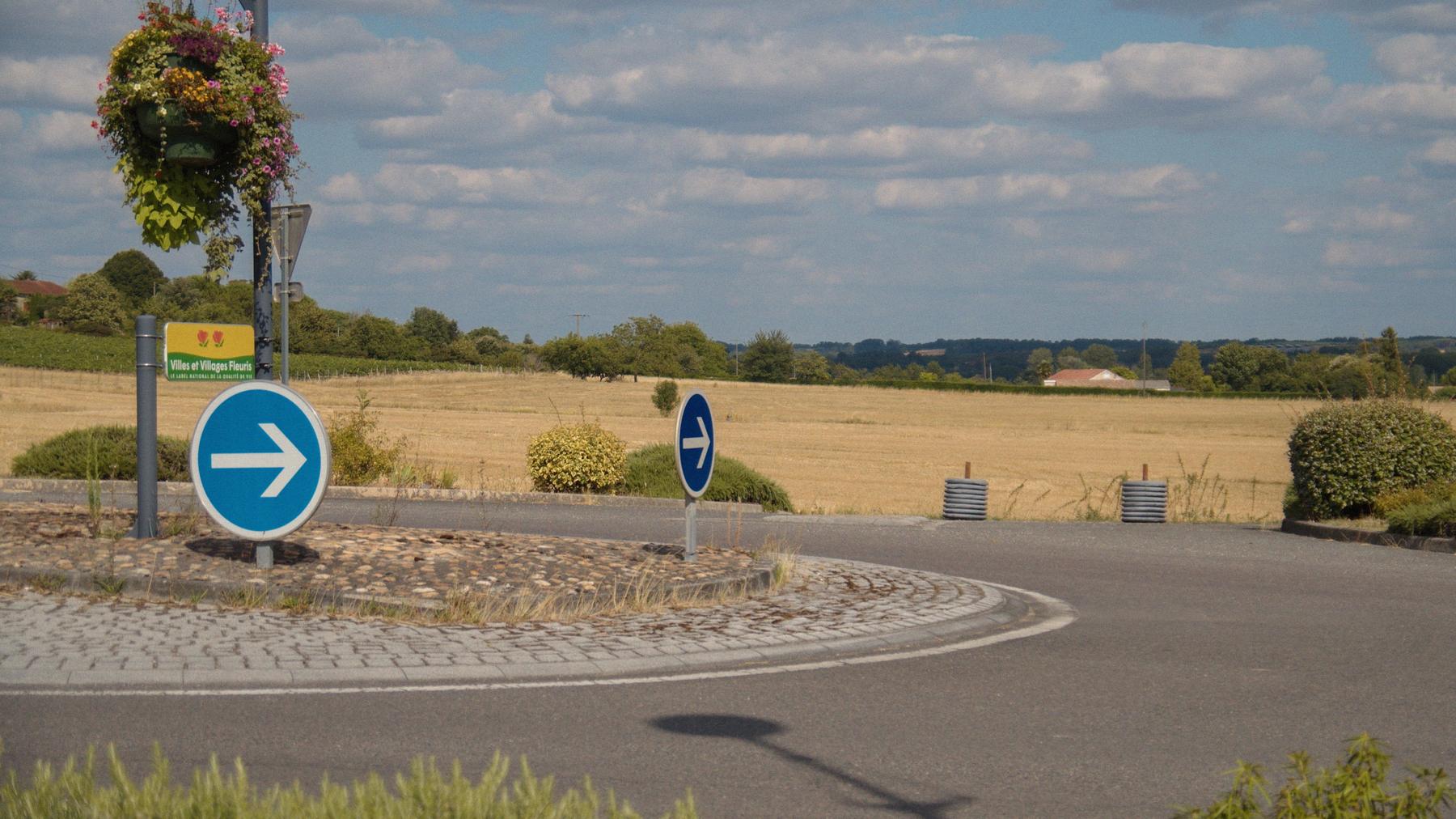 Repérage : le même rond-point fleuri, deux flèches bleues sous un ciel de petits nuages.