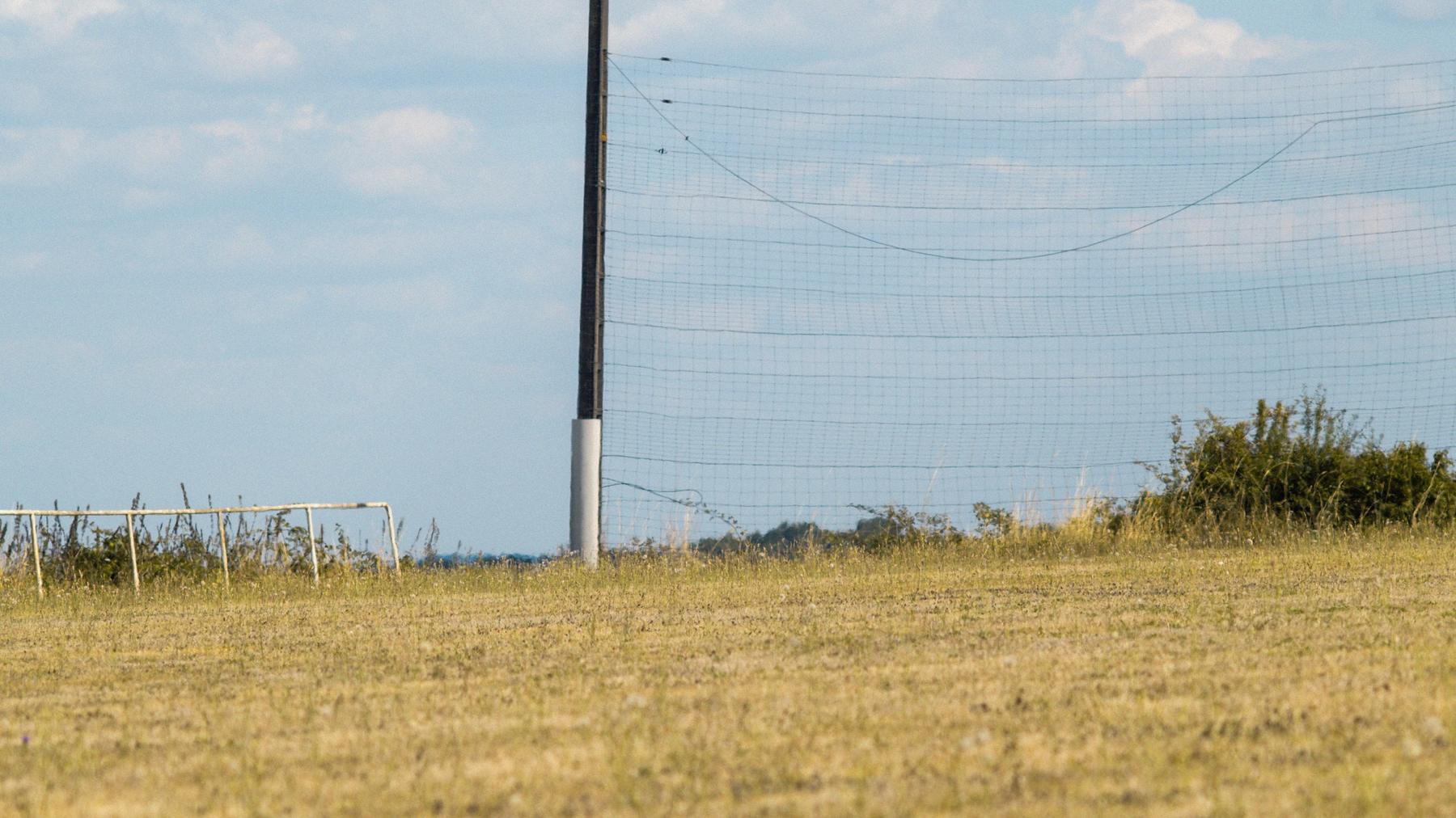 Repérage : un poteau et un filet pare-ballons au bord d'un terrain de football grillé par l'été.