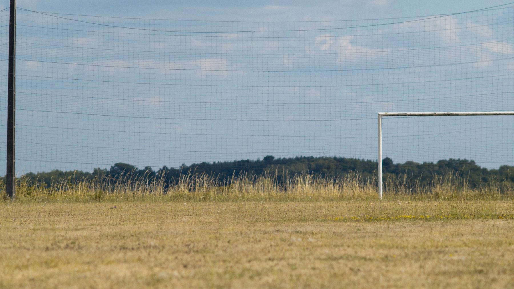 Repérage : la cage blanche d'un terrain de football de village, herbes sèches et bois sombre au fond.