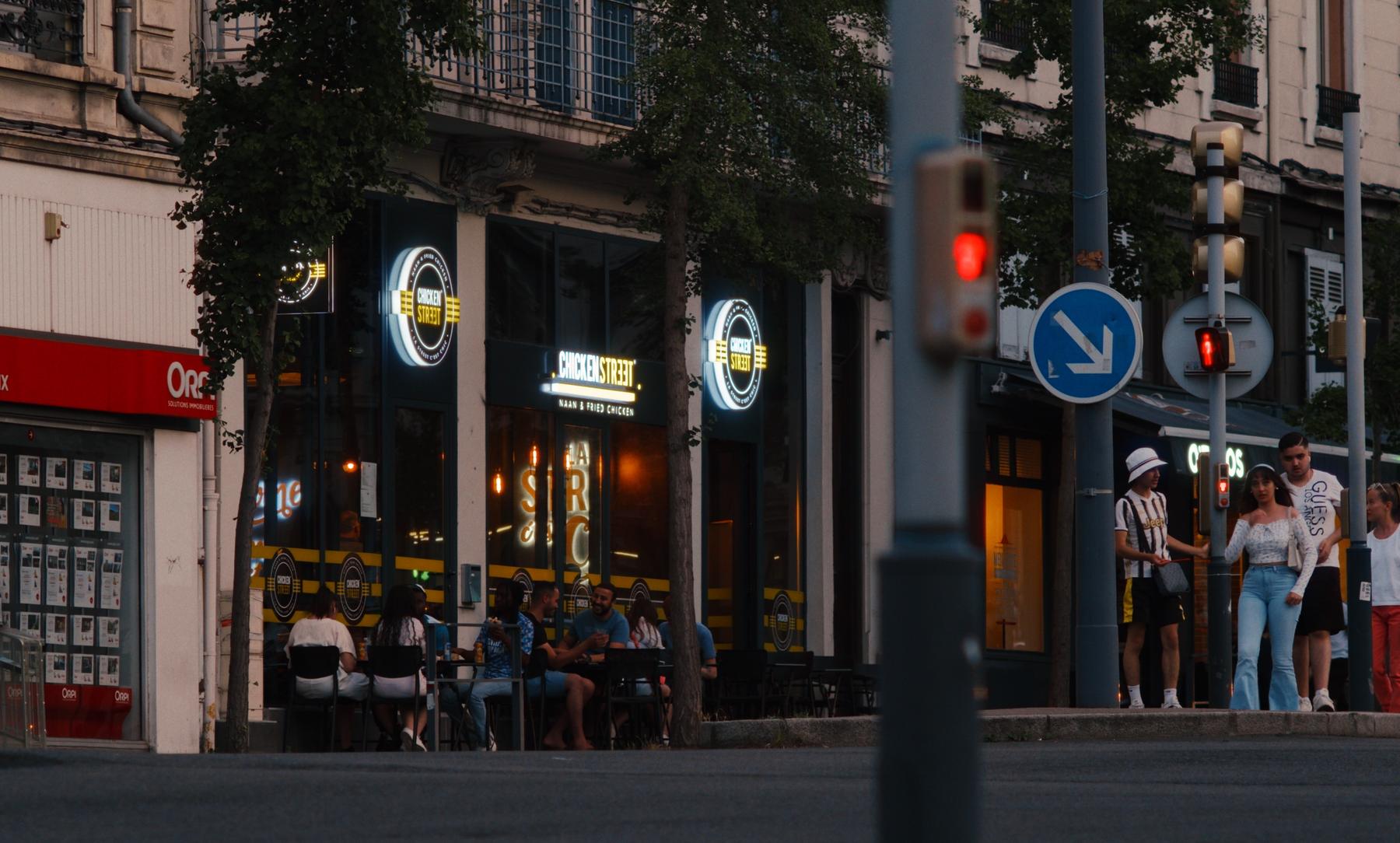 Photogramme : une terrasse de fast-food au soir, enseignes allumées.